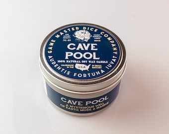 May include: A silver tin candle with a blue label that reads "Game Master Dice Company Audentis Fortuna Juvat" and "Cave Pool 100% Natural Soy Wax Candle Handmade in the U.S.A. In Small Batches". The bottom of the tin reads "Cave Pool A Mysterious Well of Earth, Moss, & Marine".