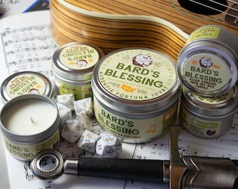 Puede incluir: Una colección de cinco latas de velas plateadas con la etiqueta "Bard's Blessing" y un diseño verde y amarillo. Las velas están rodeadas de dados blancos y un ukelele plateado.