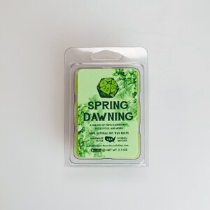 May include: A green and white soy wax melt with the text "SPRING DAWNING" and a description of the scent: "A season of fresh garden mint, eucalyptus, and herbs". The wax melt is packaged in a clear plastic container.