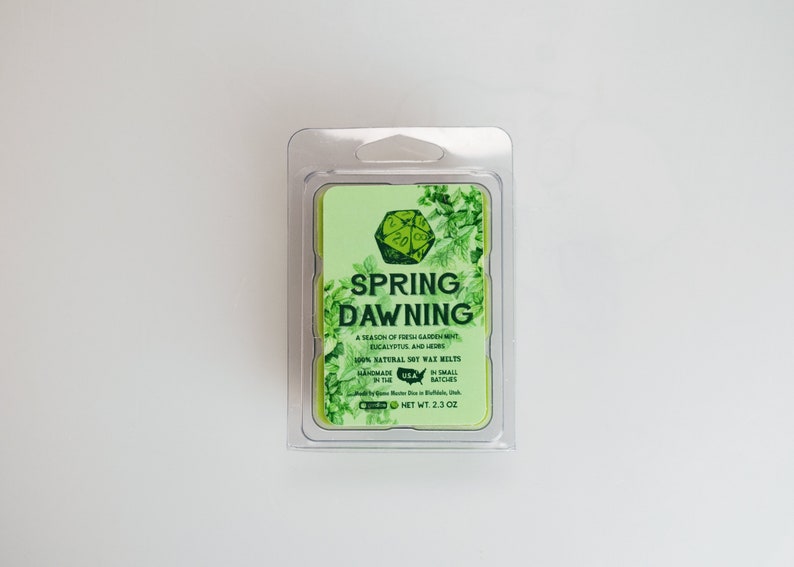 May include: A green and white soy wax melt with the text "SPRING DAWNING" and a description of the scent: "A season of fresh garden mint, eucalyptus, and herbs". The wax melt is packaged in a clear plastic container.