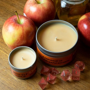 May include: Two candles in glass jars with lids, one labeled "High Elf Cider" and the other "Add a Friendship of Wine and Maple Bourbon".  The candles are surrounded by red apples and dice.