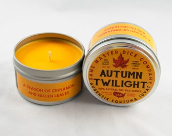 May include: Two small, round, silver candle tins with lids. The candle in the foreground has a label that reads "Game Master Dice Company Audentis Fortuna Juvat" and "Autumn Twilight 100% Natural Soy Wax Candle" in red and black text. The candle in the background has a label that reads "A Season of Cinnamon and Fallen Leaves".