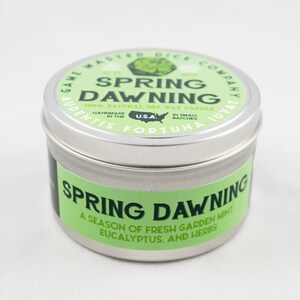 May include: A silver tin candle with a green label that reads "Spring Dawning 100% Natural Soy Wax Candle" and "A season of fresh garden mint, eucalyptus, and herbs".