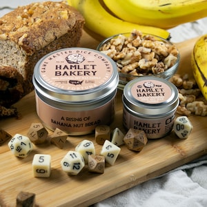 May include: Two small tin candle jars with the label "Hamlet Bakery" and the scent "A Rising of Banana Nut Bread". The candles are on a wooden surface with a bowl of walnuts, a loaf of banana nut bread, and dice.