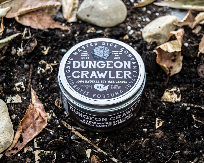 May include: A silver tin candle with a black label that reads "Game Master Dice Company" and "Dungeon Crawler". The candle is 100% natural soy wax and is handmade in the USA. The label also includes the text "Audentis Fortuna Juvat" and "Brown on American Soil".