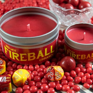 May include: Two red cinnamon scented candles in silver tins with the words "Red Hot Fireball Cinnamon" on the front. The candles are surrounded by red candy and red and yellow dice.