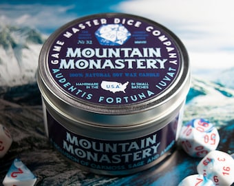 May include: A silver tin candle with a blue and purple label that reads "Game Master Dice Company" and "Mountain Monastery 100% Natural Soy Wax Candle." The label also includes the text "Audentis Fortuna Iuvat" and "Handmade in the U.S.A. In Small Batches." The candle is surrounded by blue and white dice.