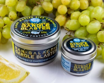 Puede incluir: Dos velas de estaño plateado con etiquetas negras. La vela más grande está etiquetada como "Pan-Galactic Gargle Blaster" y dice "A smashing of Chardonnay & Lemon". La vela más pequeña está etiquetada como "Pan-Galactic Gargle Blaster" y dice "Galaxy's Finest 100% Soy Wax Candle". Ambas velas están sobre una superficie blanca con una rodaja de limón y un racimo de uvas verdes.