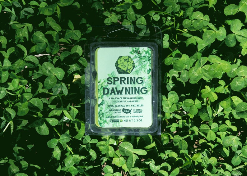 May include: A green soy wax melt in a clear plastic container with a label that reads "Spring Dawning" and describes the scent as a "season of fresh garden mint, eucalyptus and herbs". The label also states that the wax melt is 100% natural soy wax, handmade in small batches, and has a net weight of 2.3 oz.