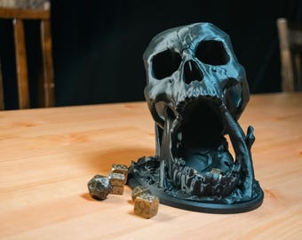 Op de afbeelding: Een zwarte 3D-geprinte schedel met een open mond, gebruikt als dobbelsteentoren. De schedel staat op een houten tafel met vier gouden dobbelstenen ernaast.