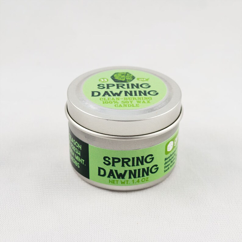 May include: A silver tin candle with a green label that reads "Spring Dawning" and "Clean-Burning 100% Soy Wax Candle". The label also has a green leaf design. The bottom of the tin reads "Net Wt. 1.4 oz".