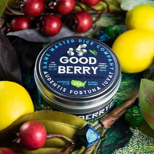 May include: A blue and silver candle tin with the text "Game Master Dice Company" and "Good Berry" printed on it. The candle is surrounded by red berries, yellow lemons, and green leaves.