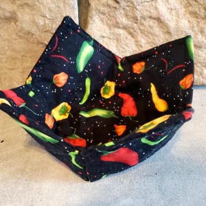 May include: A square, fabric bowl with a black background and a colorful chili pepper pattern. The peppers are red, yellow, and green, with white speckles. The bowl is open and appears to be made of quilted fabric.