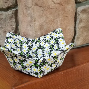 May include: A square fabric bowl with a daisy pattern. The bowl is black with white daisies and yellow centers. The bowl is sitting on a wooden surface.