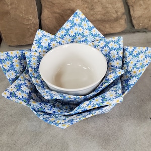 May include: A white ceramic bowl nested in a blue and white daisy-patterned fabric bowl cozy. The cozy is folded to cradle the bowl, creating a decorative and functional kitchen accessory. The background features a stone wall.