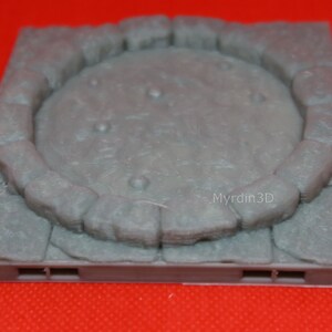 May include: A gray, textured, square-shaped tile with a raised circular stone border. The tile has a smooth, gray surface inside the border. The tile has a small, gray, rectangular base with two slots on each side.