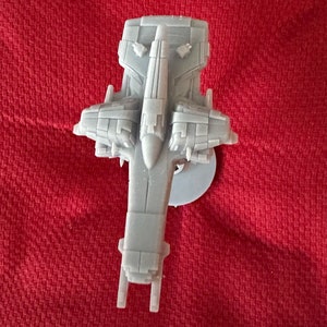 May include: A gray, miniature spaceship model with detailed features, including wings and a central body. The model is set against a red background, showcasing its intricate design and overall shape. The spaceship is likely a collectible or a display piece.