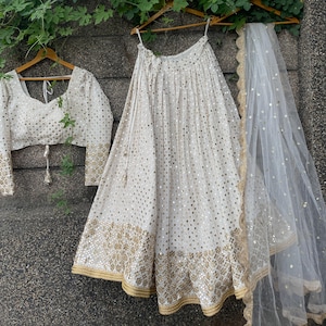 May include: A white bridal lehenga with gold embroidery and mirror work. The skirt is full-length and has a flowing silhouette. The top is a cropped style with a sweetheart neckline. The dupatta is sheer and has a delicate border.