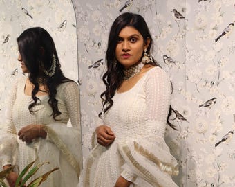 May include: A white maxi dress with a sheer overlay and gold embroidery. The dress has a round neckline and long sleeves. The dress is decorated with a gold trim and a gold tassel.