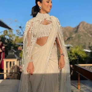 May include: A white saree with a white embroidered cape and pearl embellishments. The saree is draped over a woman's body and the cape is worn over her shoulders. The woman is standing in front of a mountain range.