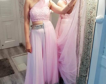May include: A pink one-shoulder crop top with white embroidery and a matching pink skirt with a silver belt. The skirt has a sheer overlay and a silver trim.