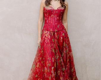 May include: A red floral print dress with a corset-style bodice and a flowing skirt. The dress features a strapless design and is adorned with a gold and red jeweled choker. The dress is suitable for formal events.
