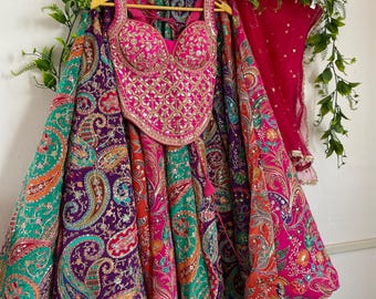 May include: A vibrant lehenga set featuring a sleeveless, sweetheart-neckline bodice heavily embellished with gold embroidery and mirrors. The voluminous, floor-length skirt displays a multi-colored paisley pattern in purple, teal, and pink.
