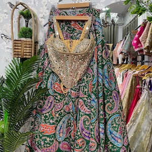 May include: A green paisley print skirt and a gold-colored, embellished corset top. The skirt features a vibrant pattern with shades of green, blue, orange, and purple. The corset is adorned with reflective embellishments.