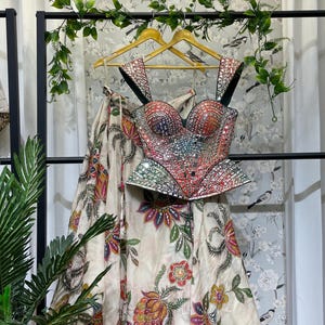 Puede incluir: Un conjunto formal que presenta un top tipo bustier y una falda. El top está adornado con detalles de espejo y presenta un escote corazón. La falda está adornada con un estampado floral en tonos rojos, naranjas y verdes. El conjunto se exhibe en una percha.