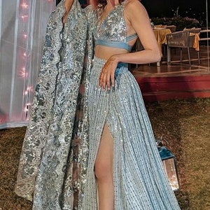 May include: A woman wearing a light blue sequined crop top and matching skirt with a high slit. The skirt has a silver and white pattern. She is wearing her hair in a bun and has pink highlights.