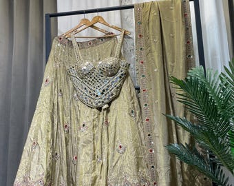 May include: A gold-colored, embellished formal outfit, including a skirt, top, and shawl, hangs on a rack. The top is heavily decorated with mirrored accents. The skirt and shawl feature intricate embroidery with red, green, and gold details. The outfit is designed for special occasions.