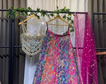 May include: A vibrant, multi-colored formal outfit, including a top with mirror embellishments and a patterned skirt. A sheer pink scarf with sequins hangs nearby. The outfit is displayed on a clothing rack.