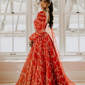 Puede incluir: Una mujer con un lehenga rojo y dorado bordado con una blusa de manga larga y hombros abullonados. Lleva una diadema y joyas doradas.