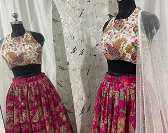 May include: A two-piece formal outfit on a mannequin. The top is a halter style with a white background and a colorful paisley pattern. The skirt is a vibrant fuchsia with a gold paisley design. A sheer white scarf is draped over the mannequin's shoulder.