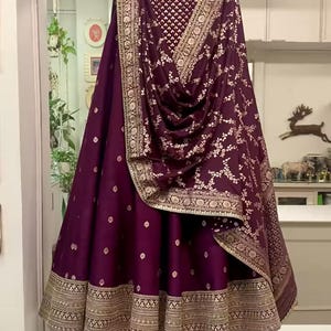 May include: A deep purple lehenga skirt and matching dupatta with gold embroidery. The skirt has a detailed gold border at the hem. The dupatta features a floral pattern and gold trim. The outfit is hanging on a wooden hanger.