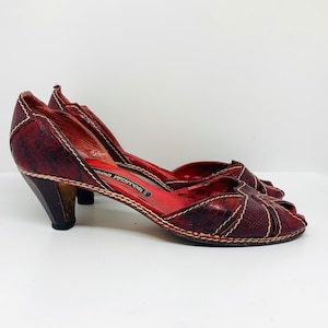 Puede incluir: Un par de tacones rojos de cuero con puntera abierta, estampado de piel de serpiente y un pequeño tacón de bloque. Los zapatos son de la marca "Ego  Pistor".