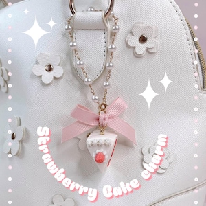 May include: A white handbag with a gold handle and floral embellishments. A charm hangs from the handle, featuring a slice of strawberry cake with a pink bow and pearl chain. The text "Strawberry Cake charm" is also visible.