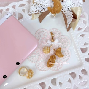 May include: A pink handheld gaming console, a gold-toned ring with a textured bread-shaped charm, and a gold-toned charm strap featuring miniature bread shapes and a white ribbon, all displayed on a white tray with a lace-patterned doily.
