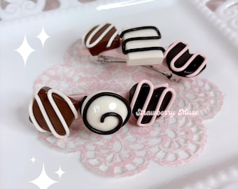 May include: Two hair clips featuring miniature chocolate-shaped charms. The top clip has three brown and white chocolate pieces with drizzle patterns. The bottom clip features three chocolate pieces with white and pink icing details. Text reads Strawberry Muse.