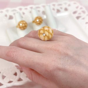 May include: A ring featuring a round, tan, textured charm resembling a melon pan bread bun with a small, clear, embedded rhinestone at its center. The charm has a grid-like pattern on its surface. The text Strawberry Muse is visible.