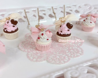 Op de afbeelding: Miniatuur cupcake bedels met roze of witte glazuur, bruine of witte vormpjes, kleine hartvormige decoraties en kleine witte parels. Elke bedel is versierd met een lichtroze strik en bevestigd aan een dunne gouden ketting. Tekst: Strawberry Muse.