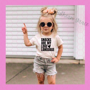 May include: A young girl wearing a white t-shirt with the text "Snacks are my love language" and a heart symbol. She is wearing denim shorts and sunglasses.
