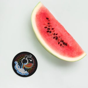 K&ouml;nnte beinhalten: Ein gestickter Patch in Schwarzwei&szlig; mit einem Regenbogen und einer Teekanne, die eine Fl&uuml;ssigkeit ausgie&szlig;t. Der Patch befindet sich auf einem wei&szlig;en Hintergrund neben einer Wassermelonen-Scheibe.