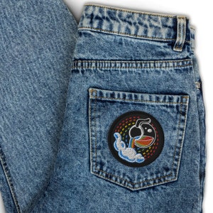 K&ouml;nnte beinhalten: Eine blaue Jeanshose mit einem schwarzen Patch auf der Ges&auml;&szlig;tasche. Der Patch zeigt einen Regenbogen, eine Wolke und eine Cartoon-Abbildung eines Kruges, der Fl&uuml;ssigkeit in eine Sch&uuml;ssel gie&szlig;t.