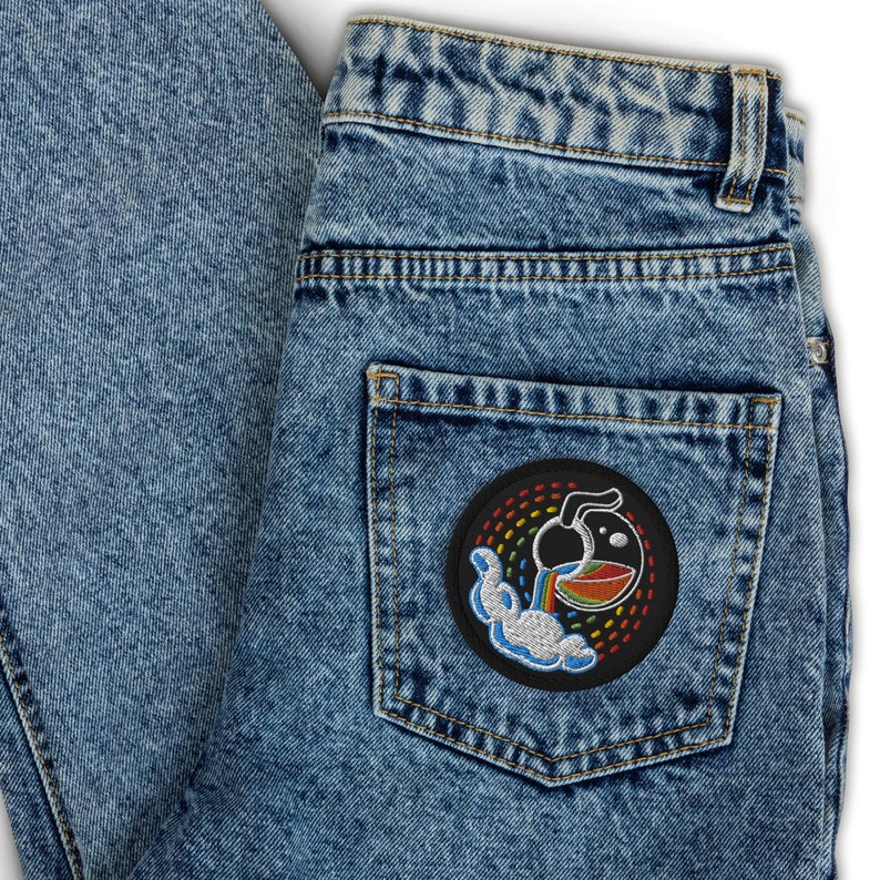 K&ouml;nnte beinhalten: Eine blaue Jeanshose mit einem schwarzen Patch auf der Ges&auml;&szlig;tasche. Der Patch zeigt einen Regenbogen, eine Wolke und eine Cartoon-Abbildung eines Kruges, der Fl&uuml;ssigkeit in eine Sch&uuml;ssel gie&szlig;t.