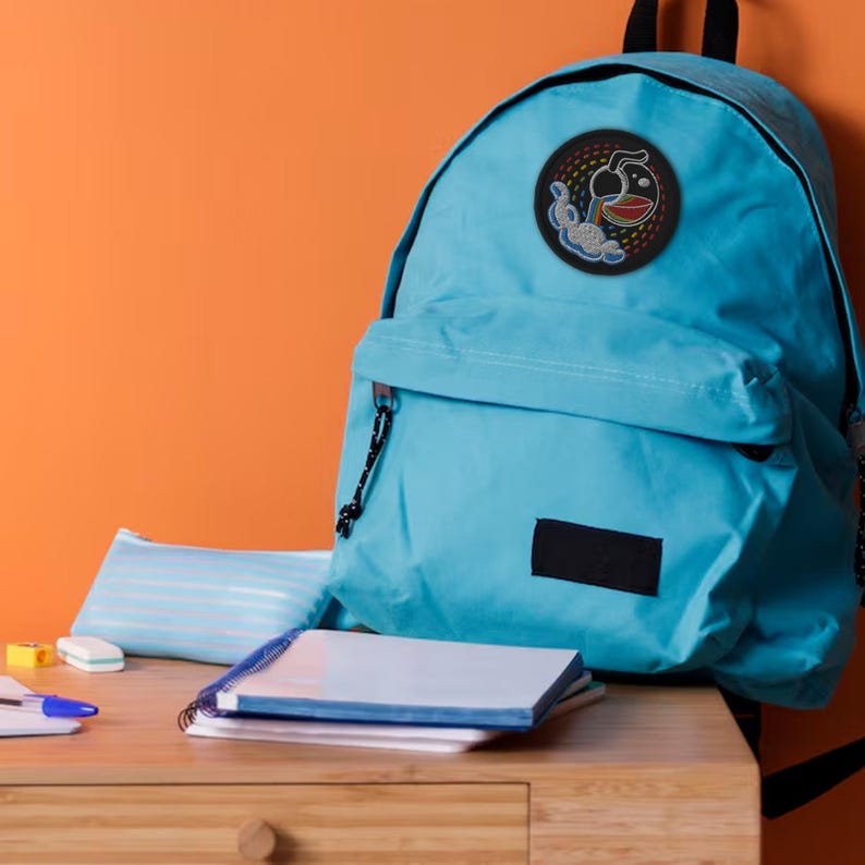 K&ouml;nnte beinhalten: Ein blauer Rucksack mit einem schwarzen und regenbogenfarbenen gestickten Aufn&auml;her eines Comic-Charakters mit einem Regenbogen und einer Wolke. Der Rucksack steht auf einem Holztisch mit einem Notizbuch und einem Federm&auml;ppchen.