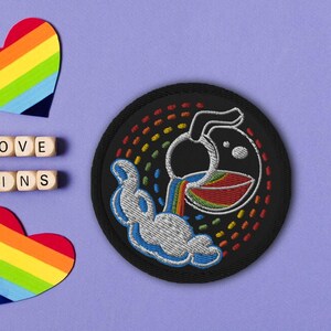 Könnte beinhalten: Ein schwarzer gestickter Patch mit einem Regenbogenmuster. Der Patch zeigt ein Cartoon-artiges Bild eines Kruges, der einen Regenbogen aus einem Krug in eine Schüssel gießt. Die Worte "LOVE WINS" sind im Hintergrund sichtbar.