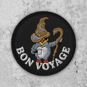 May include: A black and white embroidered patch featuring a cartoon penguin wearing a brown wizard hat and holding a staff. The text "Bon Voyage" is embroidered in white around the penguin.