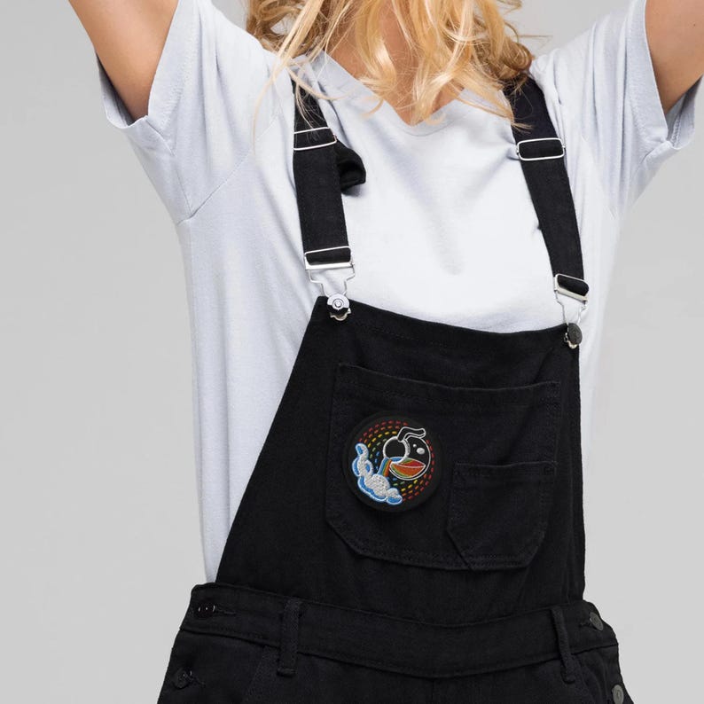 K&ouml;nnte beinhalten: Schwarze Latzhose mit verstellbaren Tr&auml;gern und einem kleinen gestickten Aufn&auml;her mit einer Cartoonfigur mit einer Bombe und einem Regenbogen.