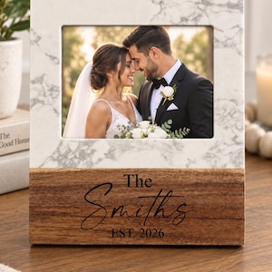 May include: A rectangular photo frame featuring a white marble top section and a natural wood base. The wood base is engraved with the text "The Smiths" in a script font and "EST. 2026" in a serif font. The frame holds a photograph of a couple.